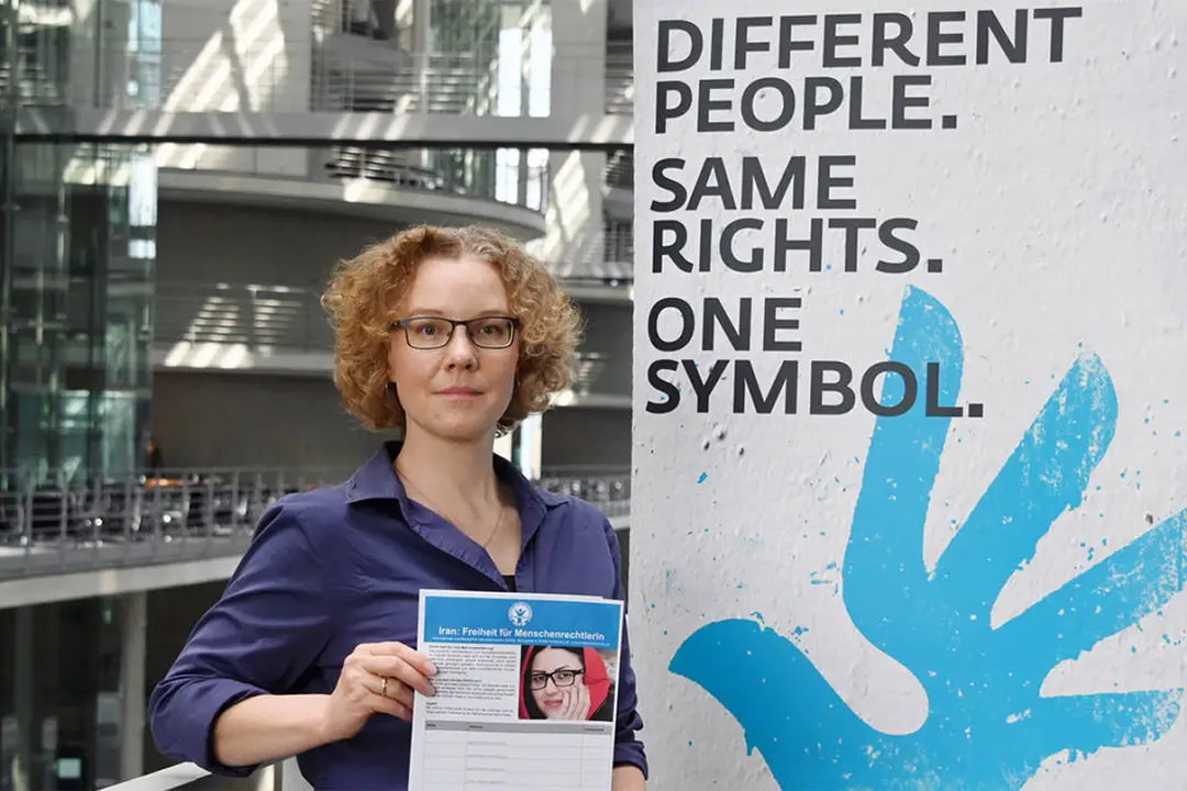 Julia Verlinden steht in einem modernen Gebäude und hält einen Zettel in die Kamera. Auf einem Plakat neben ihr steht der Slogan „Different people. Same rights. One symbol.“